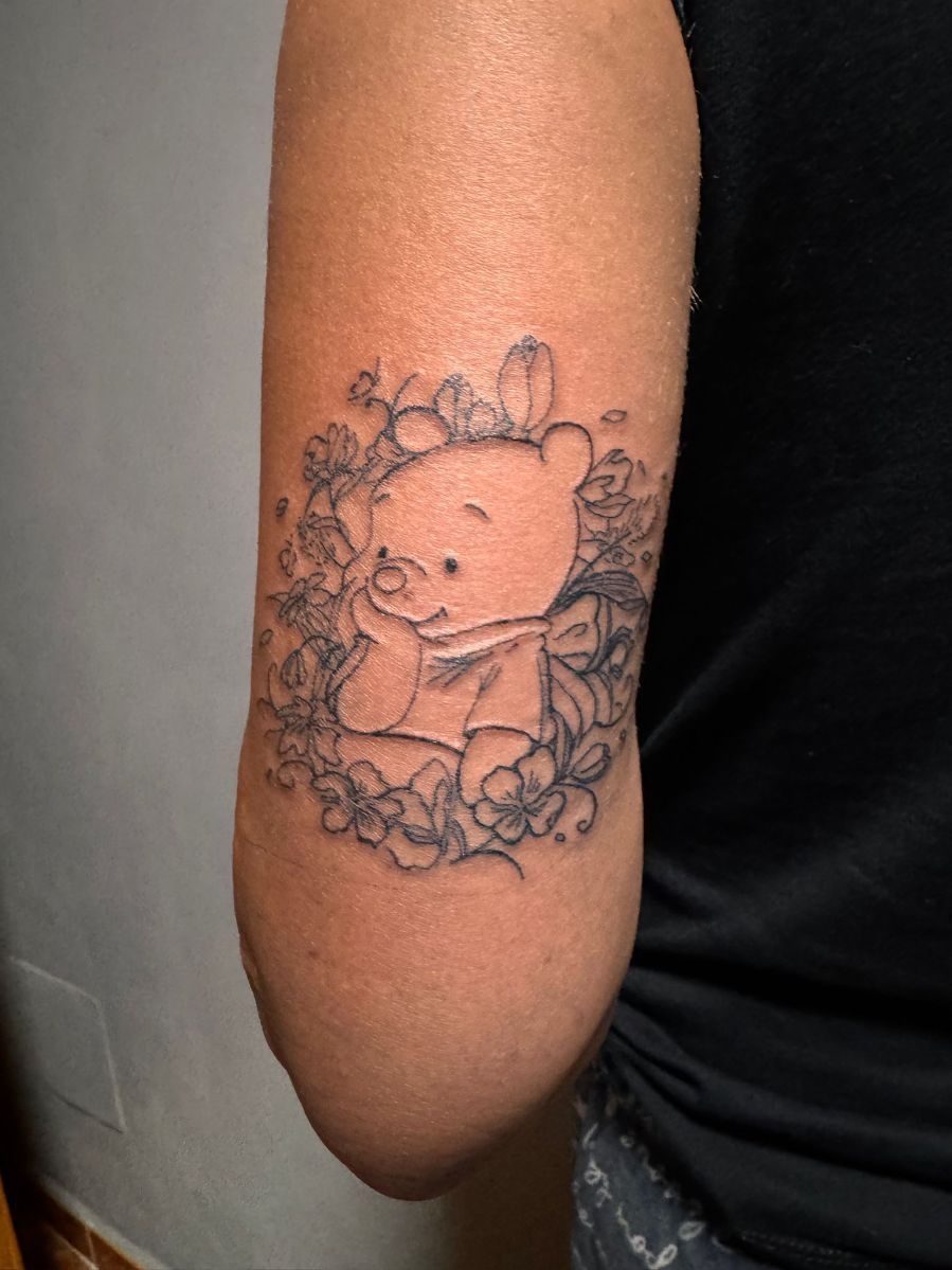 Un tatuaje de líneas finas en la parte superior del brazo de Winnie the Pooh, con la barbilla apoyada en la mano
