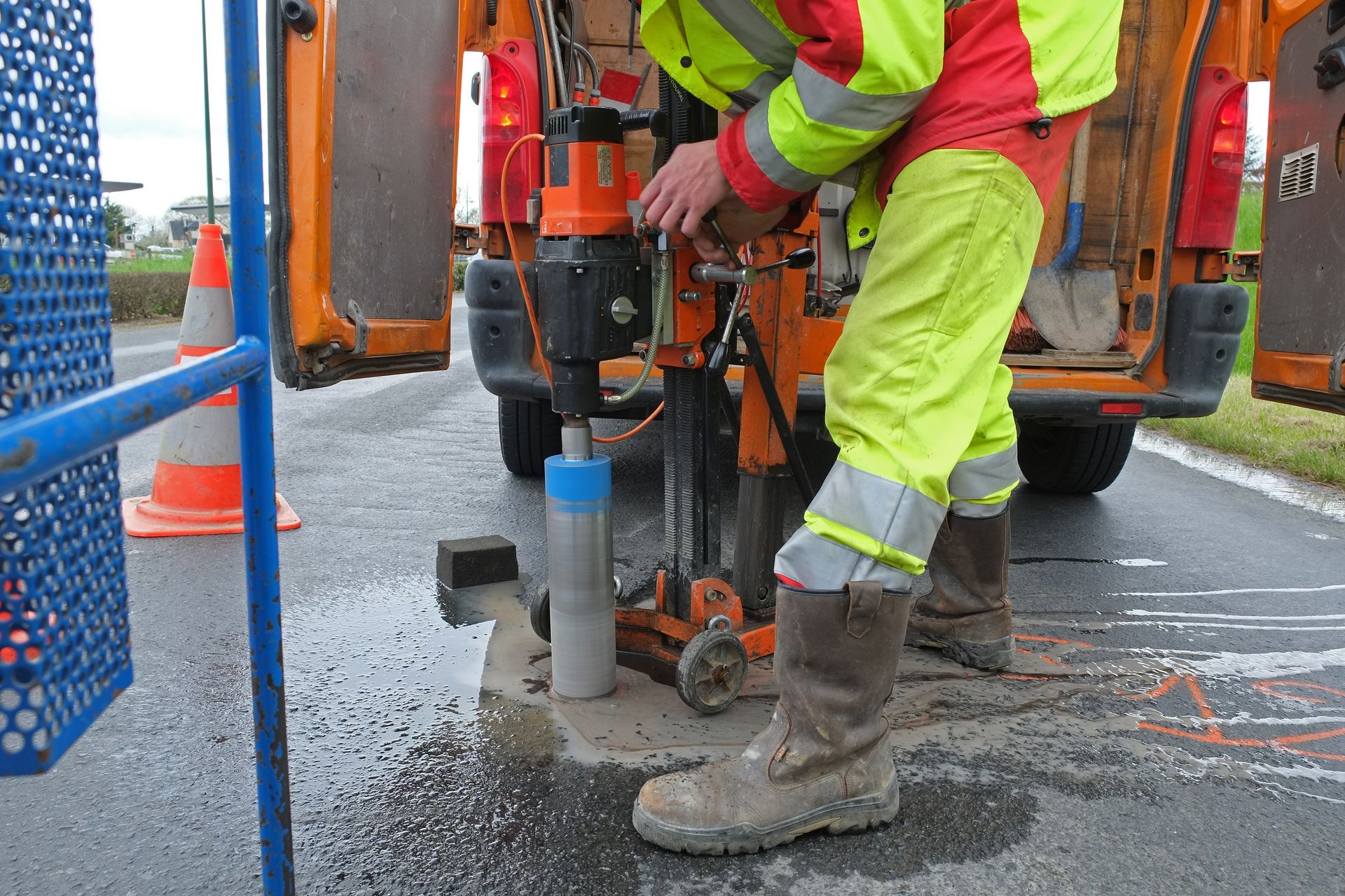 Une personne portant un équipement de sécurité utilise une carotteuse sur une route, à proximité d'un véhicule et de cônes de signalisation.