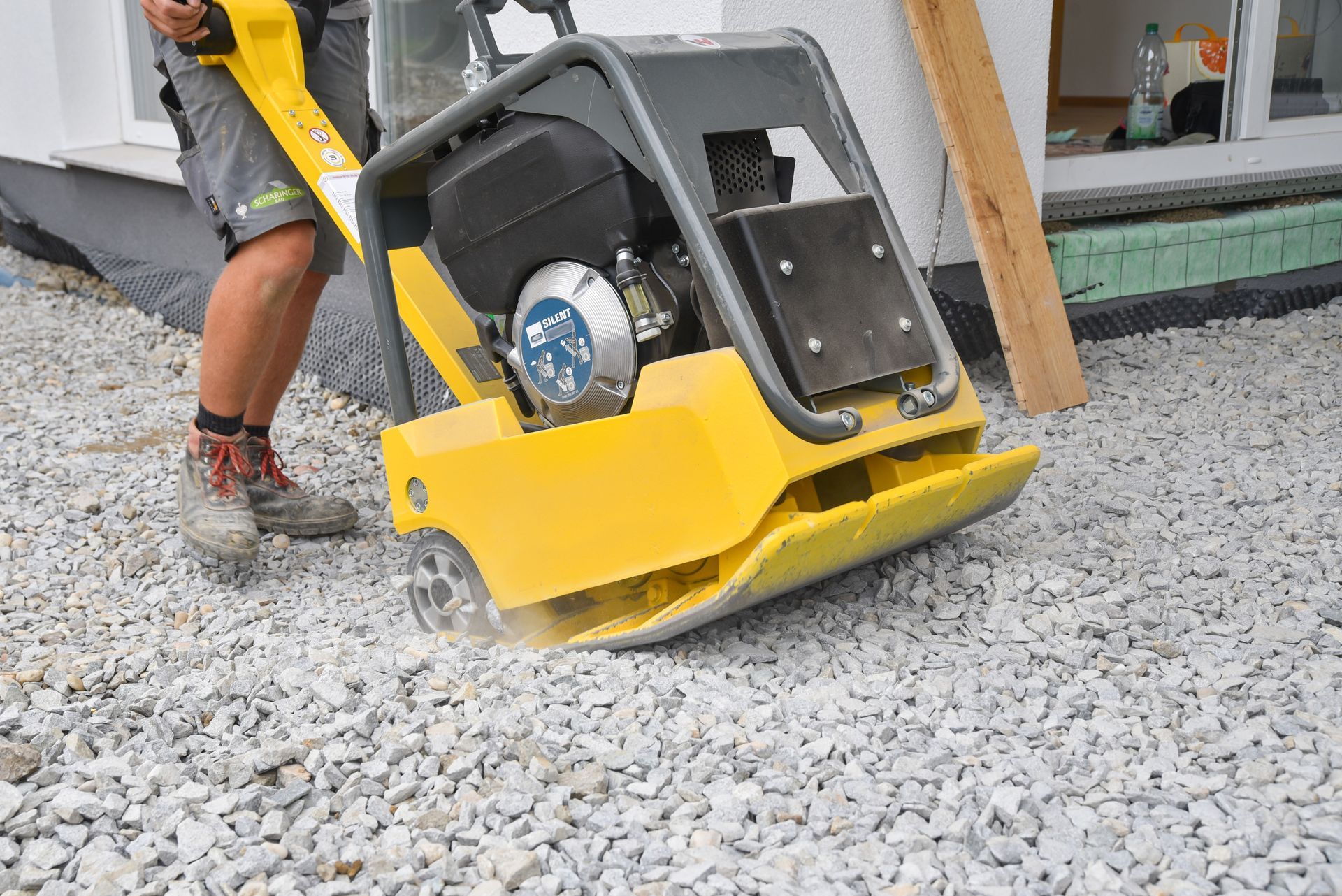 Une personne utilise une plaque vibrante jaune sur du gravier. Vue extérieure d'une maison.