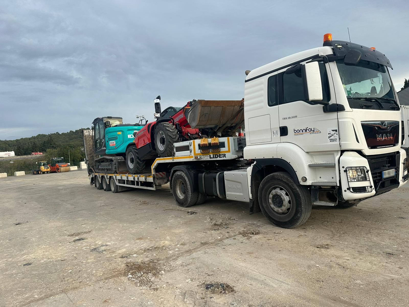 Un semi-remorque blanc transporte deux engins de chantier sur une remorque à plateau, sur un terrain gravillonné.