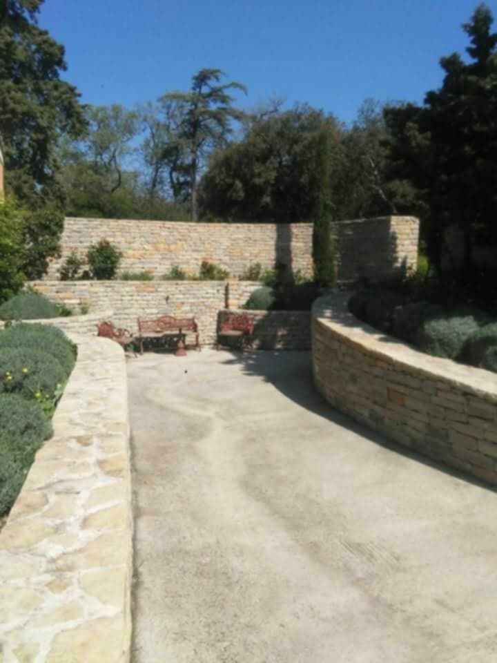 Murs en pierre et bancs dans un jardin avec un chemin en béton et un ciel bleu.