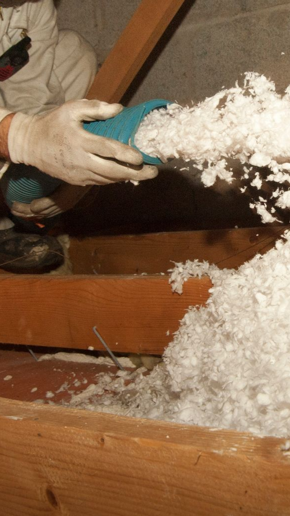 Main gantée tenant un tuyau soufflant un isolant blanc autour des poutres en bois