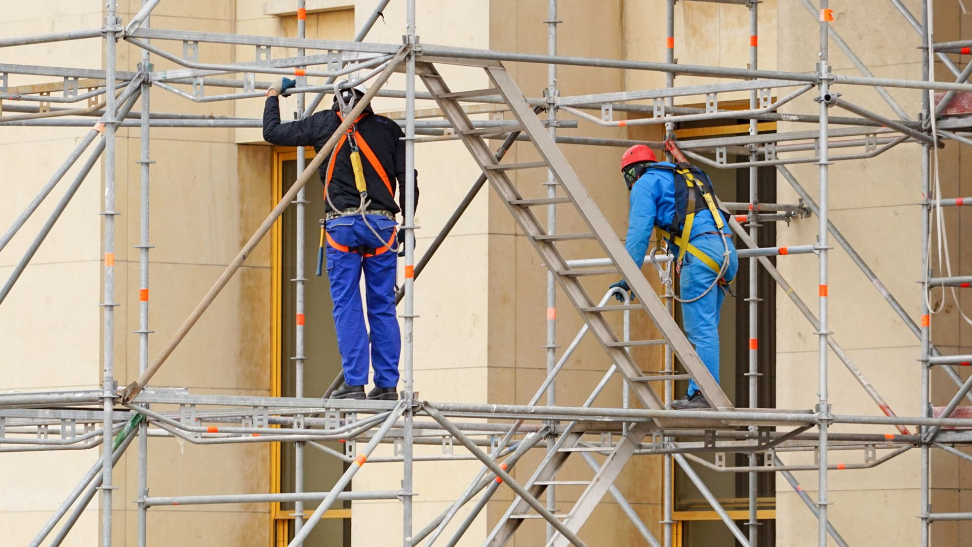 Deux ouvriers sur un échafaudage peignent la façade d'un bâtiment, l'un en salopette bleue et l'autre en casque rouge