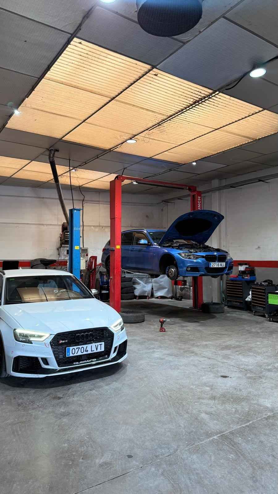 Taller mecánico con un coche blanco delante y un coche azul elevado en un elevador bajo luces brillantes del techo.
