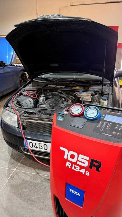 El capó del coche está abierto en un garaje, con una máquina de recarga de aire acondicionado R134a y los indicadores junto al motor.