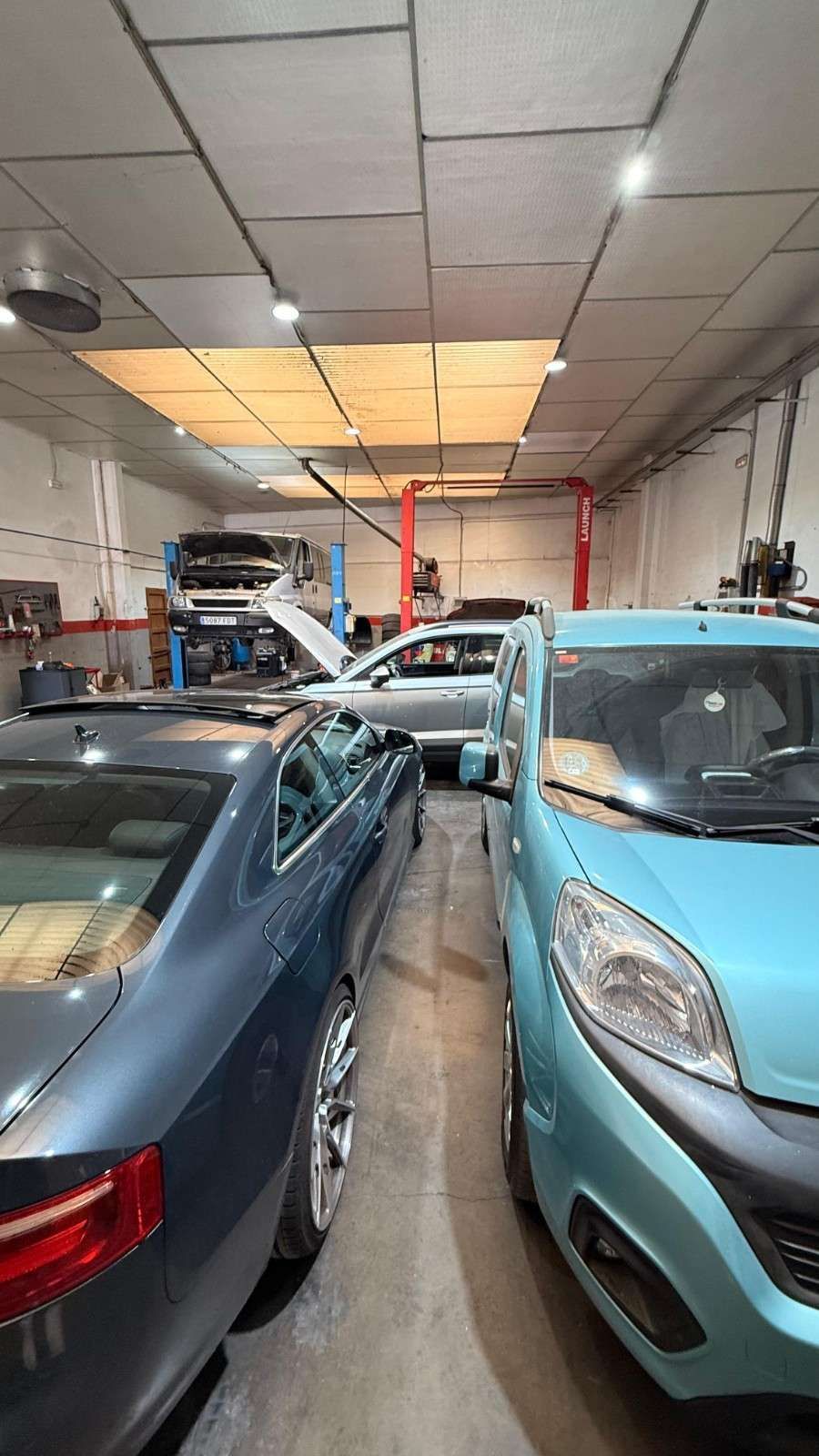Taller mecánico con un coche blanco delante y un coche azul elevado en un elevador bajo luces brillantes del techo.