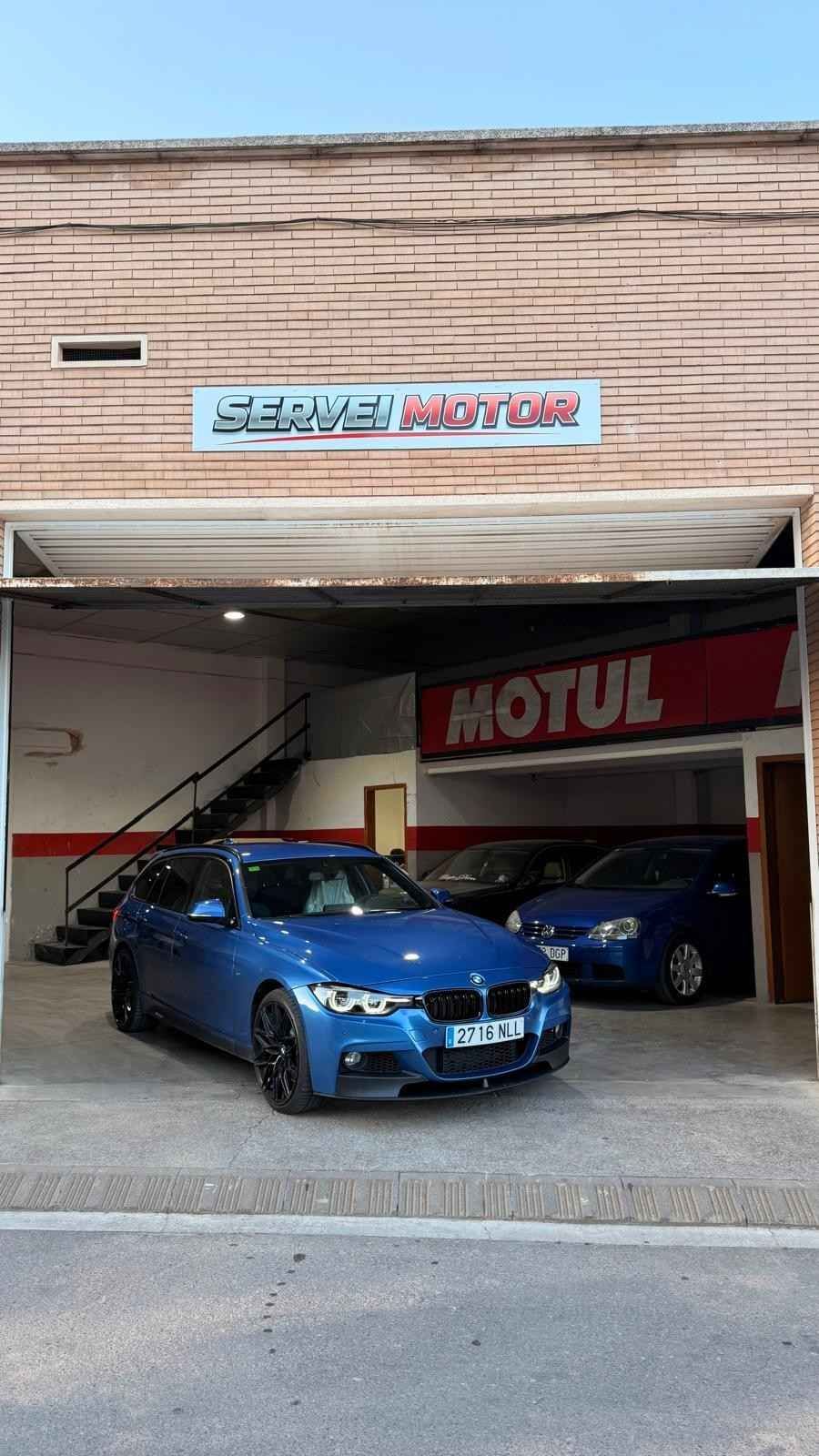 Taller mecánico con un coche blanco delante y un coche azul elevado en un elevador bajo luces brillantes del techo.