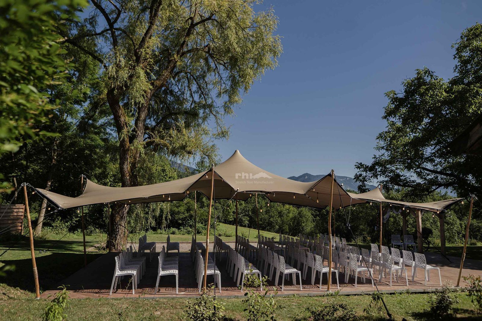 Installation d'une cérémonie de mariage en extérieur avec une tente stretch beige et des chaises blanches.
