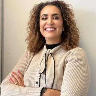 Eine Person mit lockigem Haar, die mit verschränkten Armen lächelt und eine beige Strickjacke mit dekorativer Krawatte trägt, vor einer schlichten Wand.