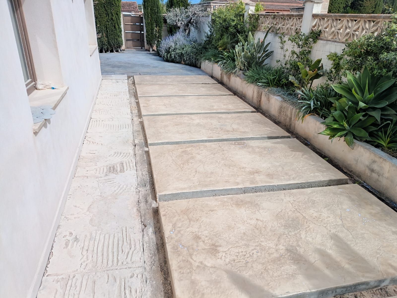 Un estrecho sendero exterior con grandes losas de piedra junto a una pared blanca y un borde de jardín plantado.