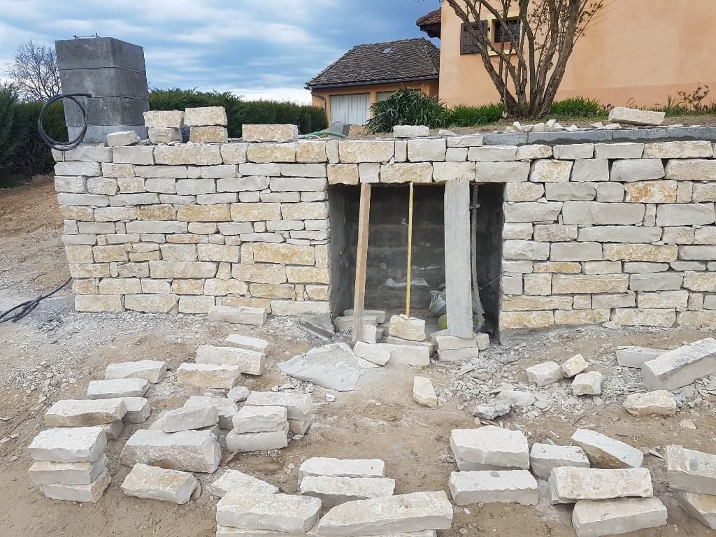 Un mur de soutènement en pierre beige partiellement construit, avec une ouverture rectangulaire et des pierres éparses au sol.