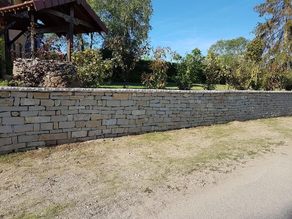 Un muret en pierre, constitué de blocs rectangulaires clairs superposés, sépare un chemin de gravier d'un jardin.