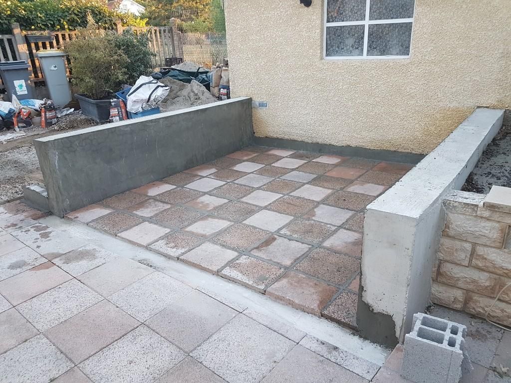 Une terrasse nouvellement construite avec un damier de dalles beiges et grises, bordée de murs de soutènement en béton.