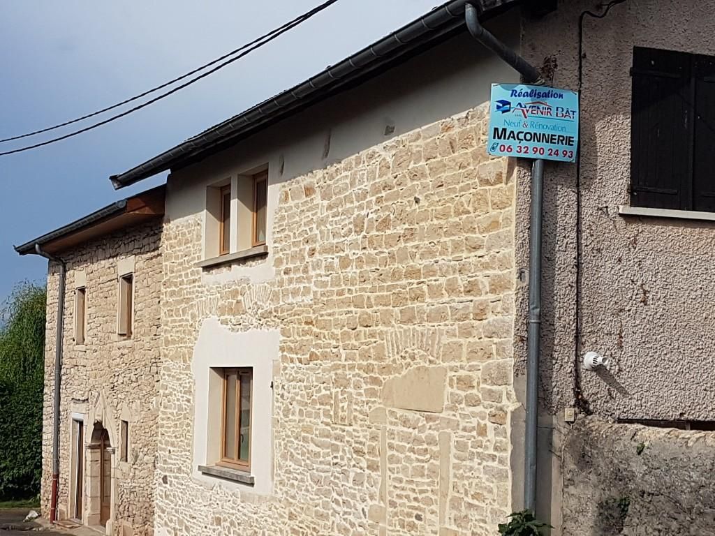 Un bâtiment en pierre d'un étage avec une façade claire, des encadrements de fenêtres en bois et un panneau publicitaire fixé au mur.