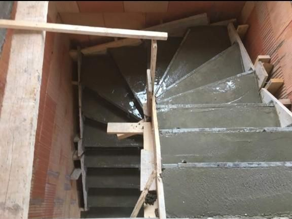 Vue de dessus d'un escalier en béton en construction entre des murs de briques, avec un coffrage en bois visible.