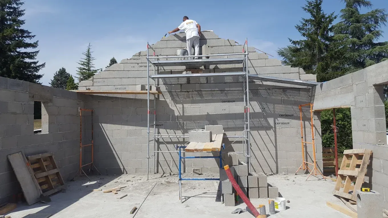 Un ouvrier du bâtiment, perché sur un échafaudage, construit un mur pignon triangulaire en béton par une journée ensoleillée.