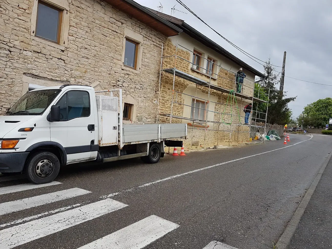 Un camion utilitaire blanc est stationné sur le bord d'une route près d'un bâtiment en pierre en cours de rénovation, présence d'un échafaudage métallique.