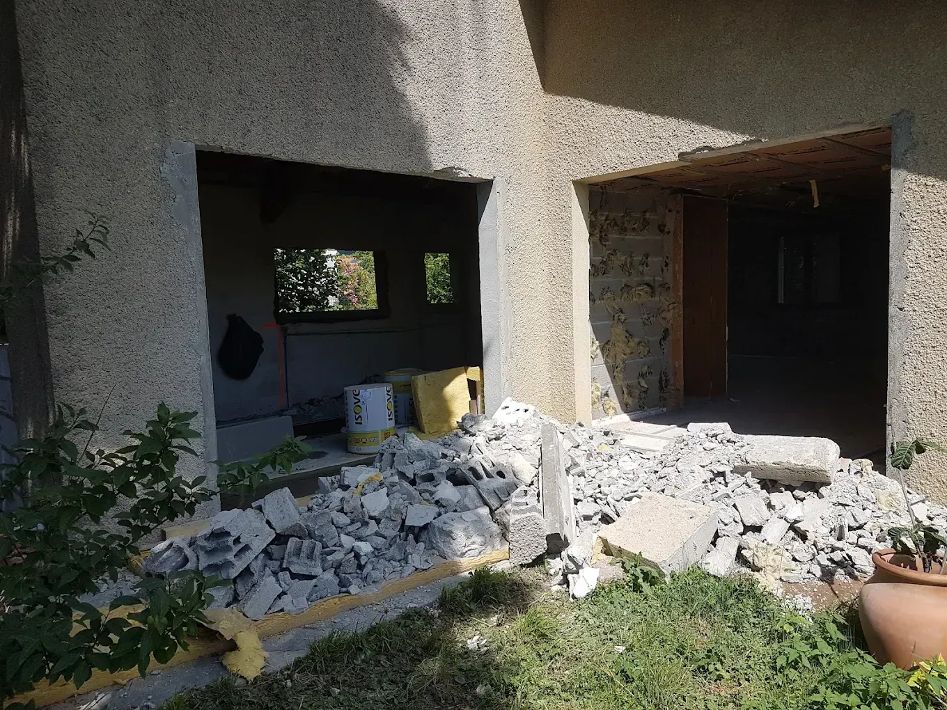 Des débris de construction jonchent le sol devant deux grandes sections de mur ouvertes, sur la façade d'une maison recouverte d'un enduit à la chaux.