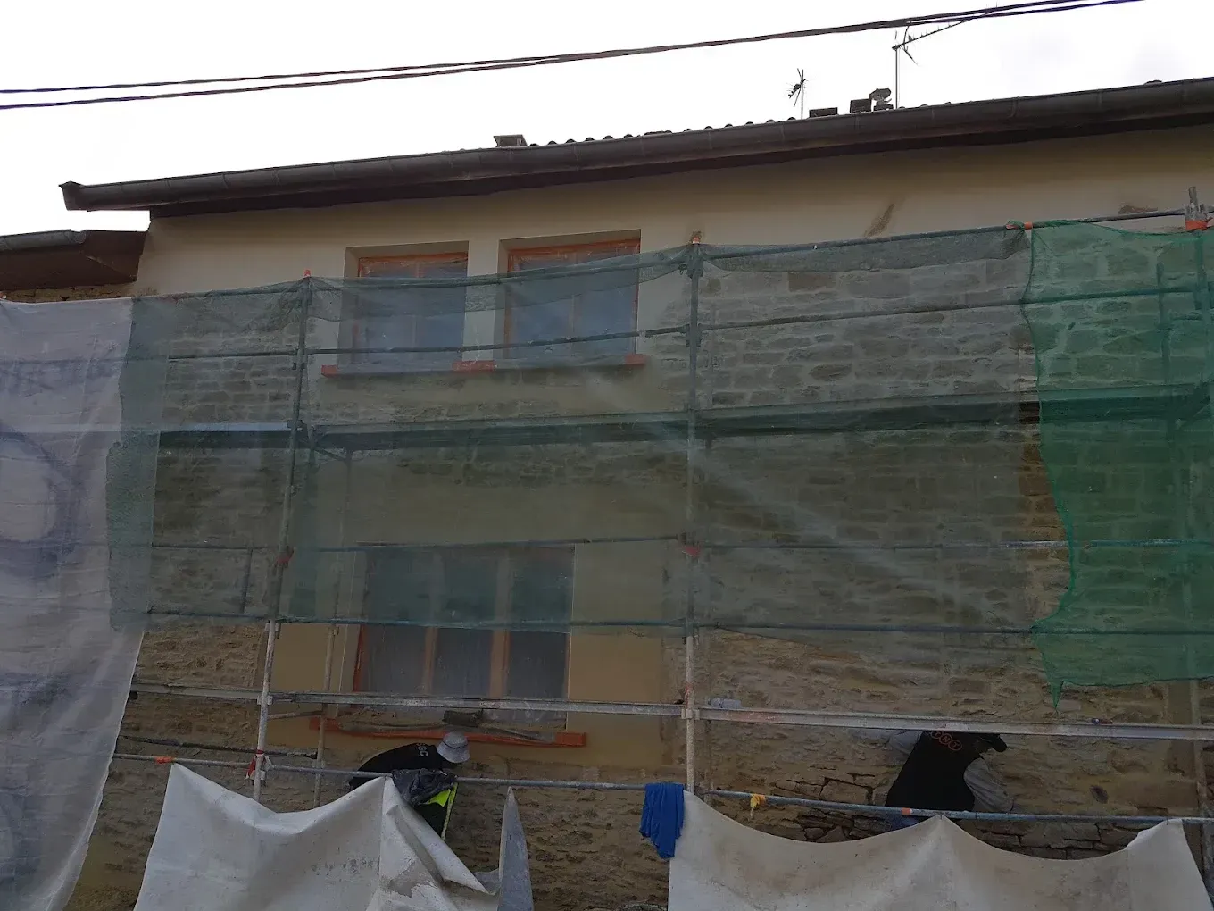 Un bâtiment en pierre claire en cours de rénovation extérieure, dissimulé par des filets de sécurité verts et des échafaudages métalliques.