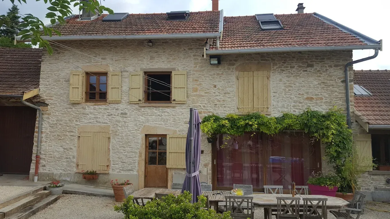 Une maison en pierre de deux étages avec un toit de tuiles, des volets en bois et un patio avec une table à manger et des parasols.