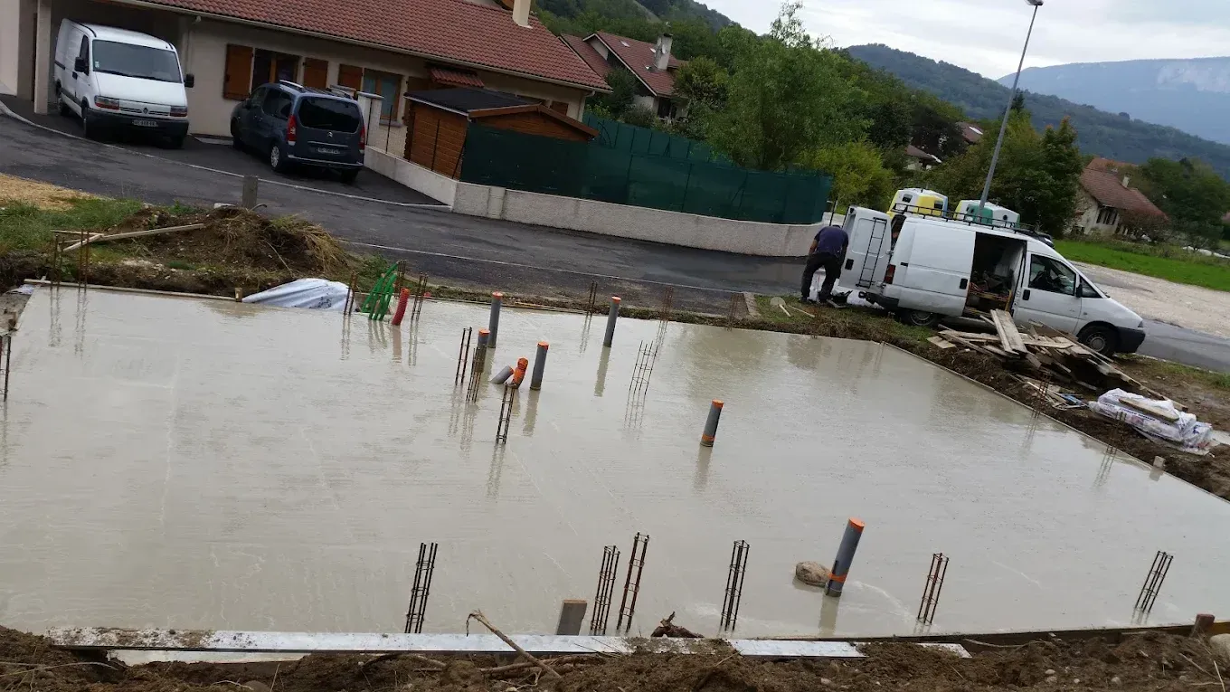 Une dalle de béton fraîchement coulée, servant de fondation à une maison, avec des tuyaux de plomberie apparents, à côté d'une camionnette utilitaire garée.