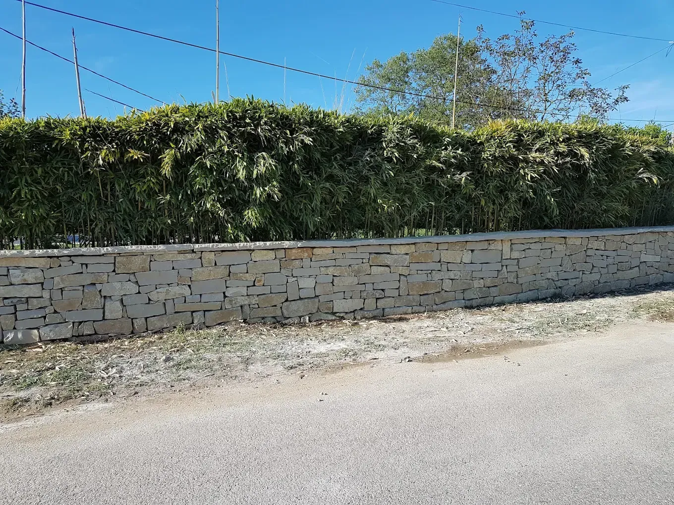 Mur de soutènement en pierre bordant un chemin de gravier, avec des haies vertes denses et un ciel bleu au-dessus.