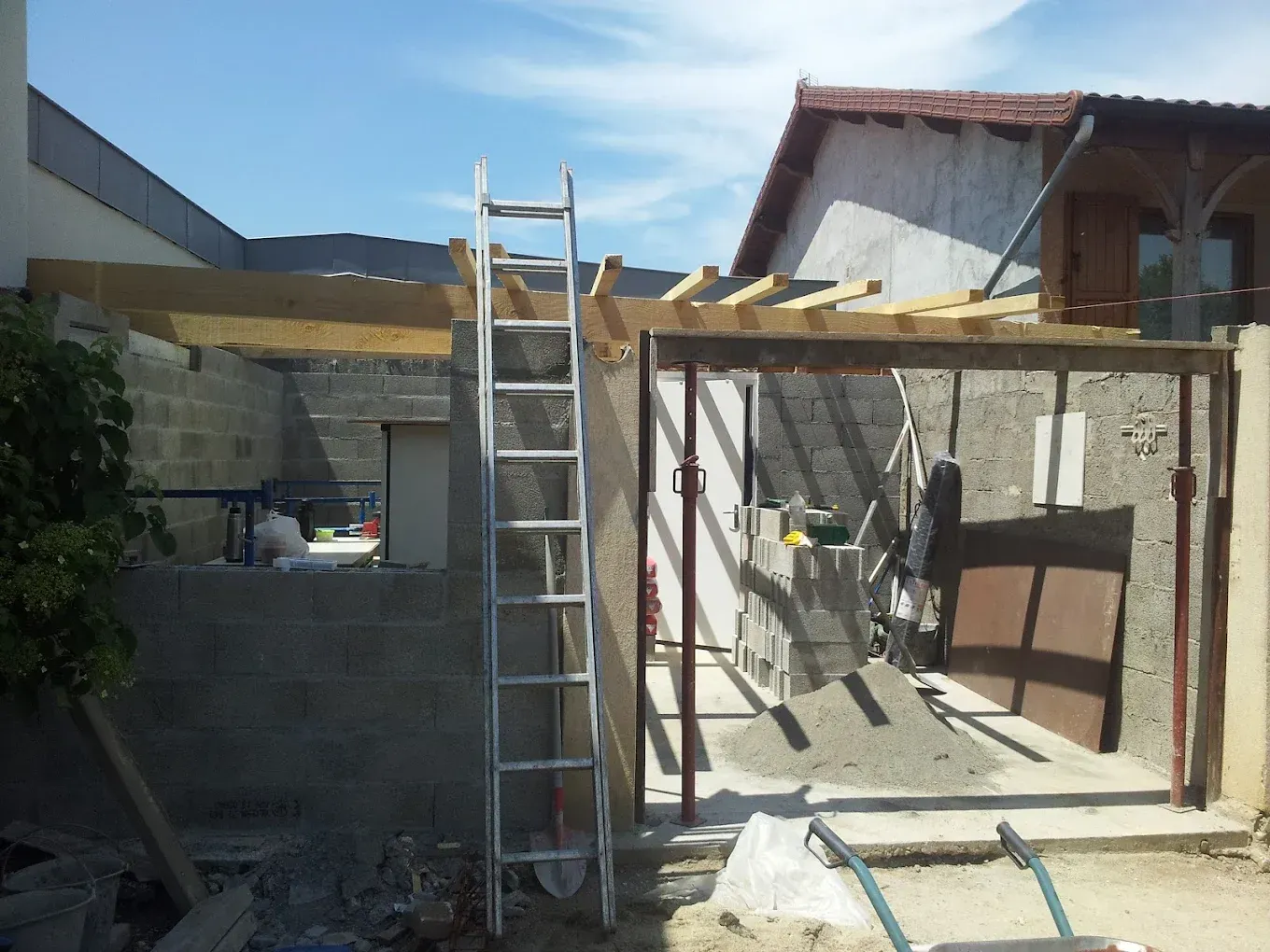 Un chantier de construction avec une structure de poutres en bois, une échelle et des poteaux de soutien, adossé à un mur en béton inachevé.