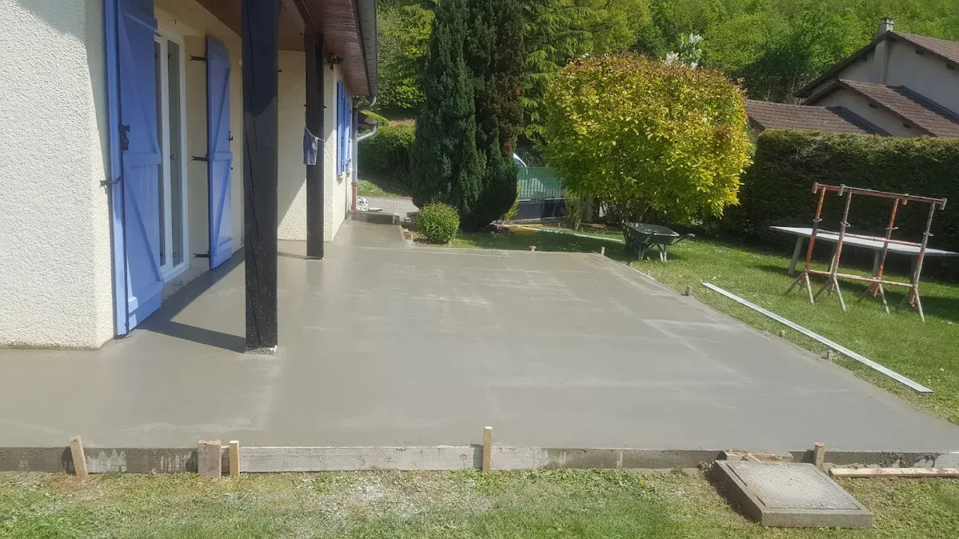 Terrasse de maison en béton gris, portes bleues et jardin gazonné avec des arbres.