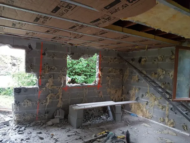 Une pièce en rénovation avec des murs en parpaings, des cadres de fenêtres ouverts et de l'isolant apparent au plafond.