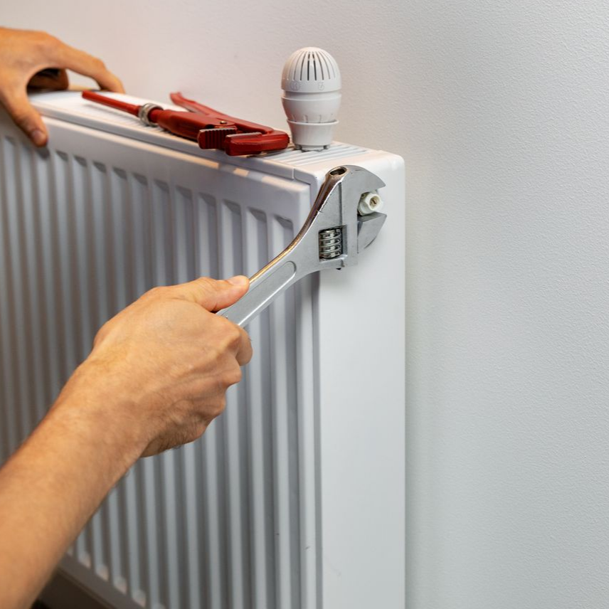 Des mains utilisent une clé pour régler une vanne de radiateur avec un outil rouge placé dessus.