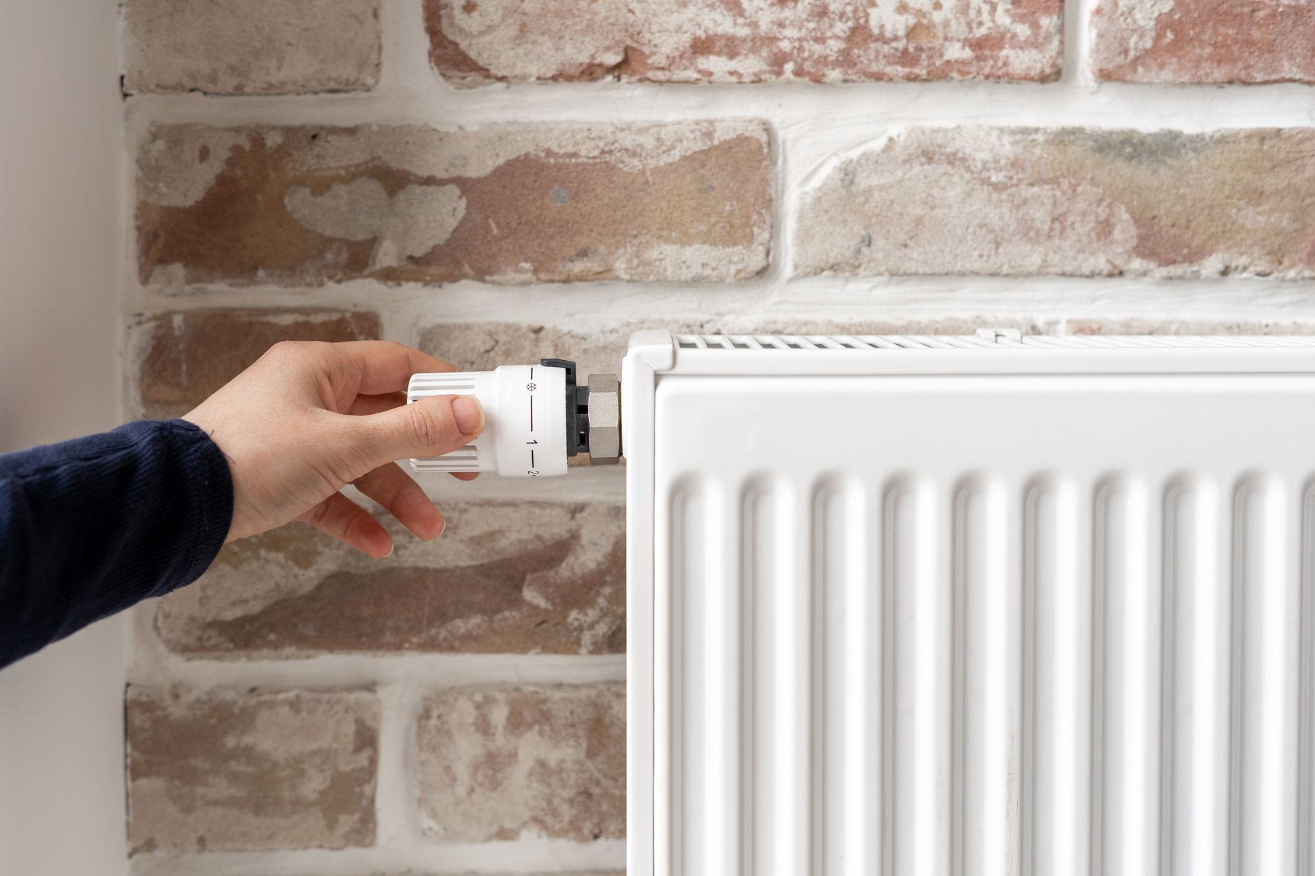Une main ajuste un thermostat à côté d'un radiateur blanc sur un mur de briques