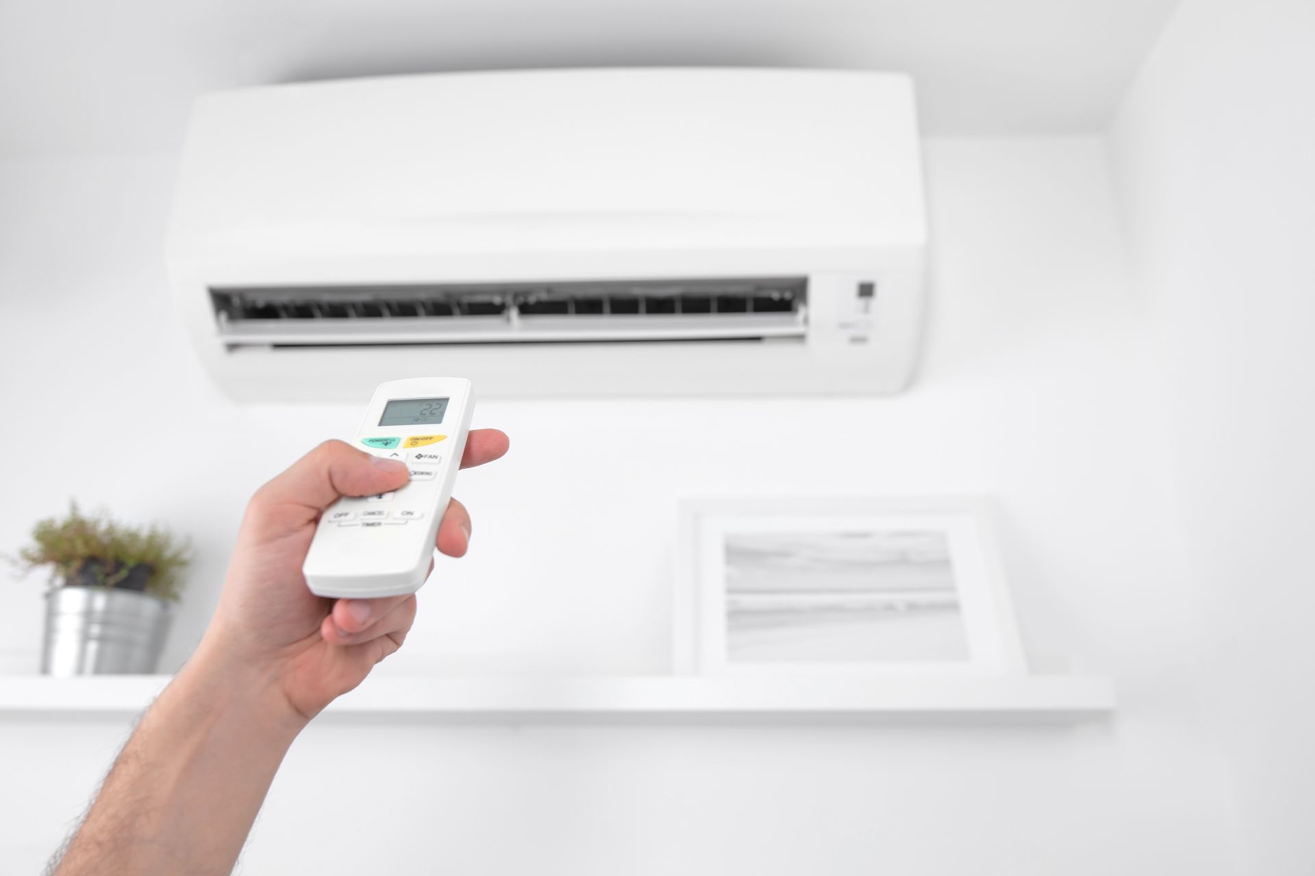 Main tenant une télécommande pointée vers un climatiseur mural blanc dans une pièce lumineuse