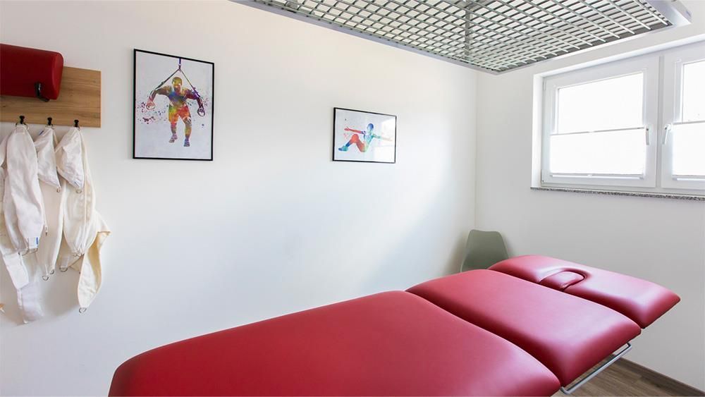 In einem Raum neben einem Fenster steht eine rote Massageliege.