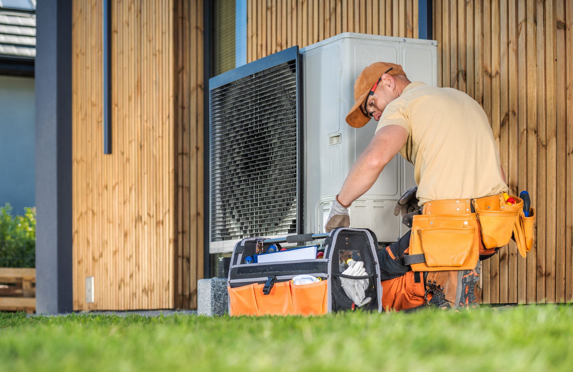 Technicien travaillant sur une unité extérieure, outils en main, sur une pelouse à côté d'un mur en bois.