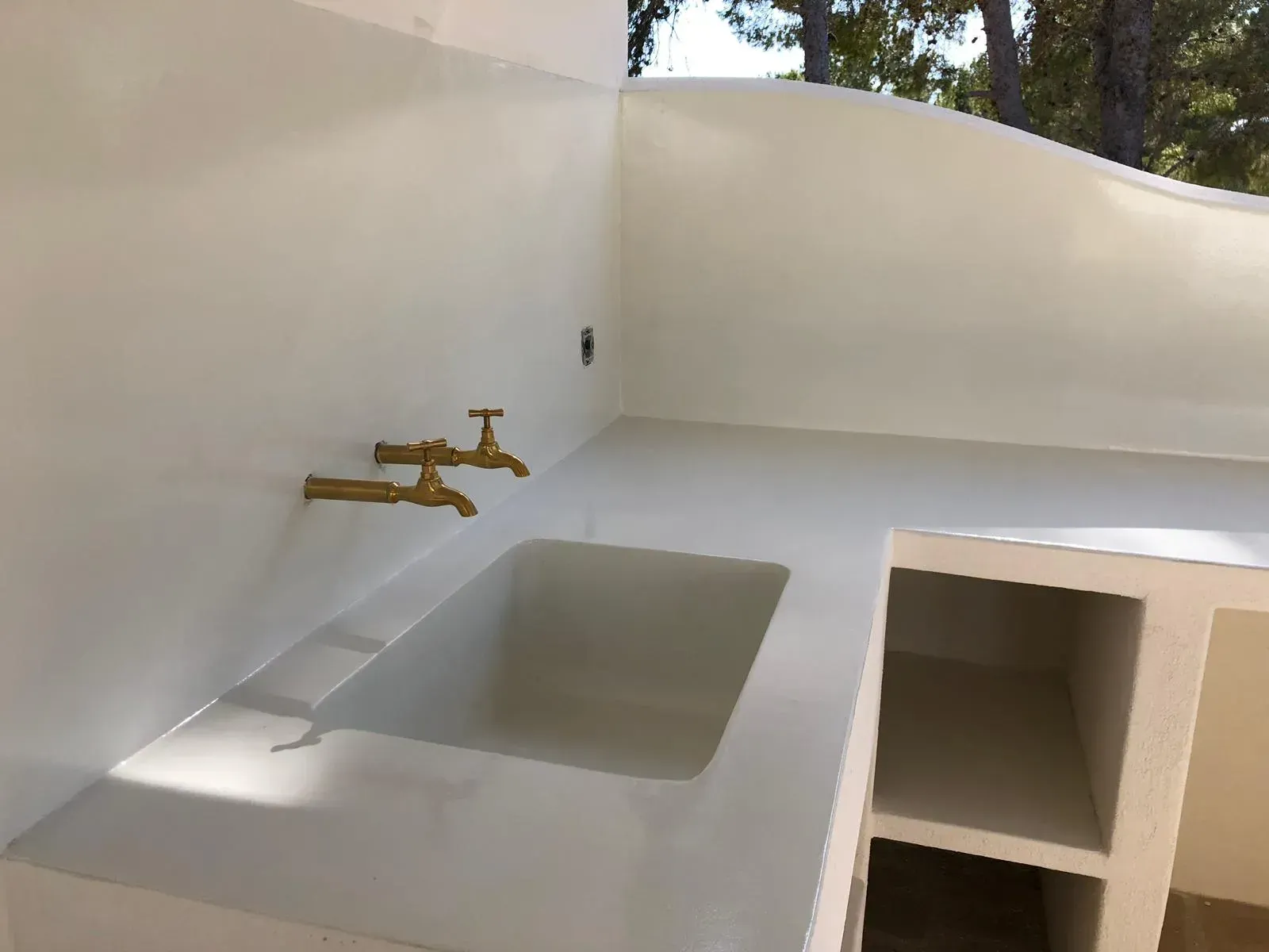 Fregadero de exterior blanco con grifería de latón, empotrado en pared, con estantes.