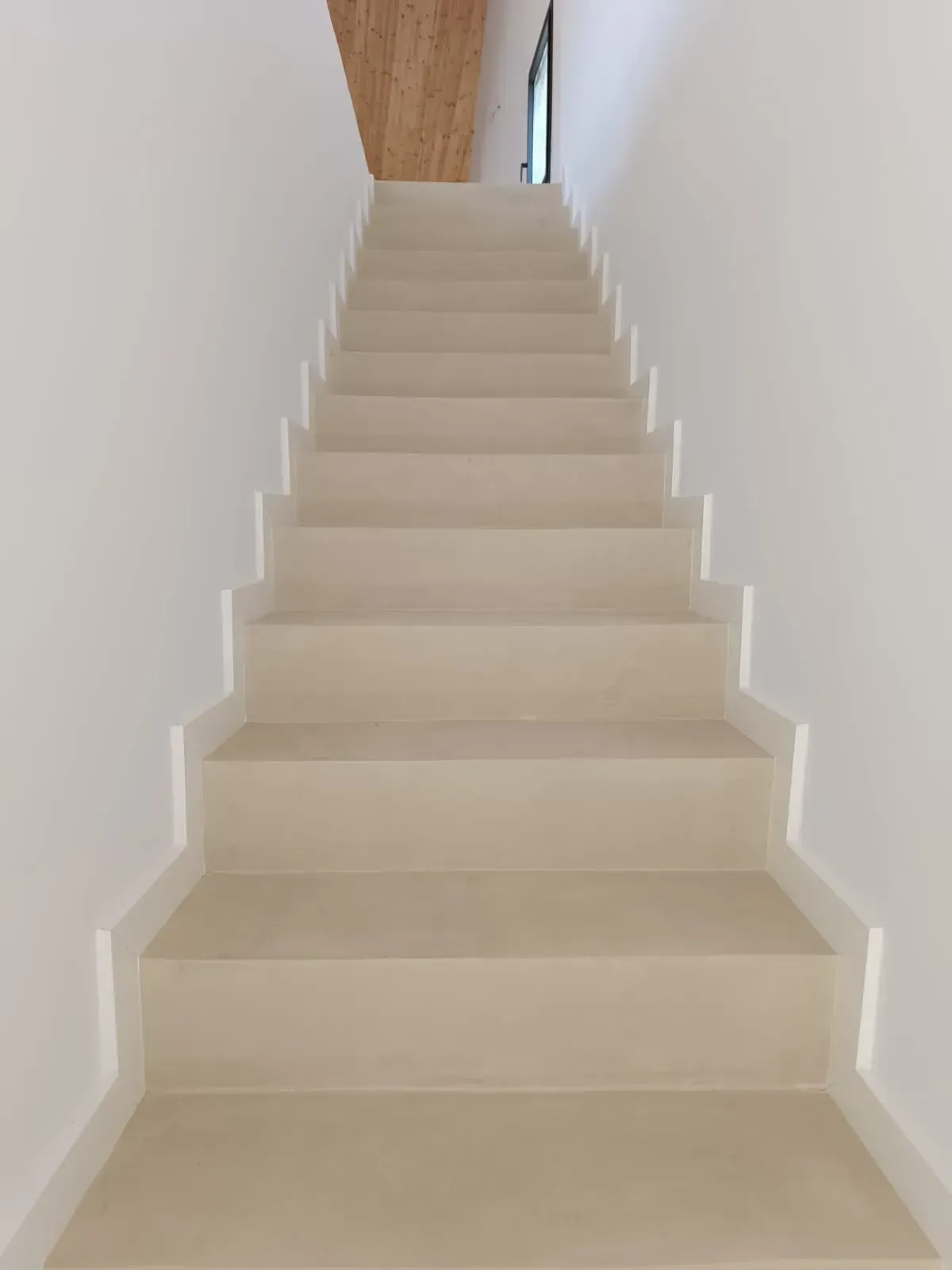 Escalera con escalones de color beige claro y paredes blancas, que conduce hacia arriba hacia un techo de madera.