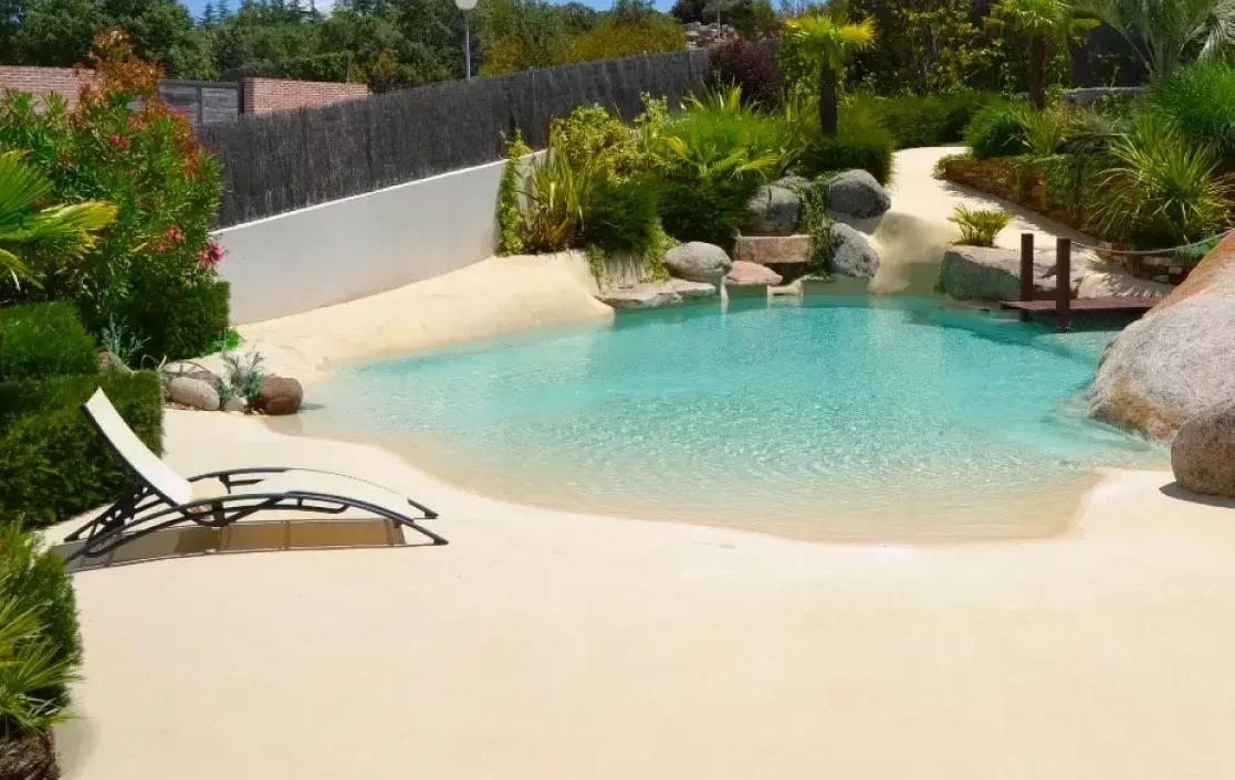 Piscina con entrada a la playa, de arena clara y agua turquesa cristalina; hay una tumbona cerca.