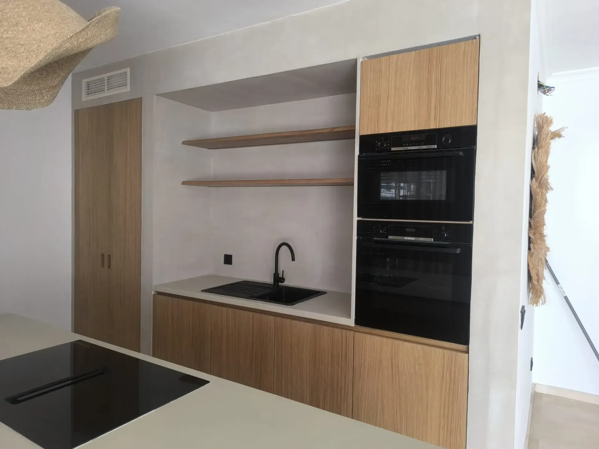 Cocina moderna con gabinetes de madera clara, hornos empotrados y fregadero negro.