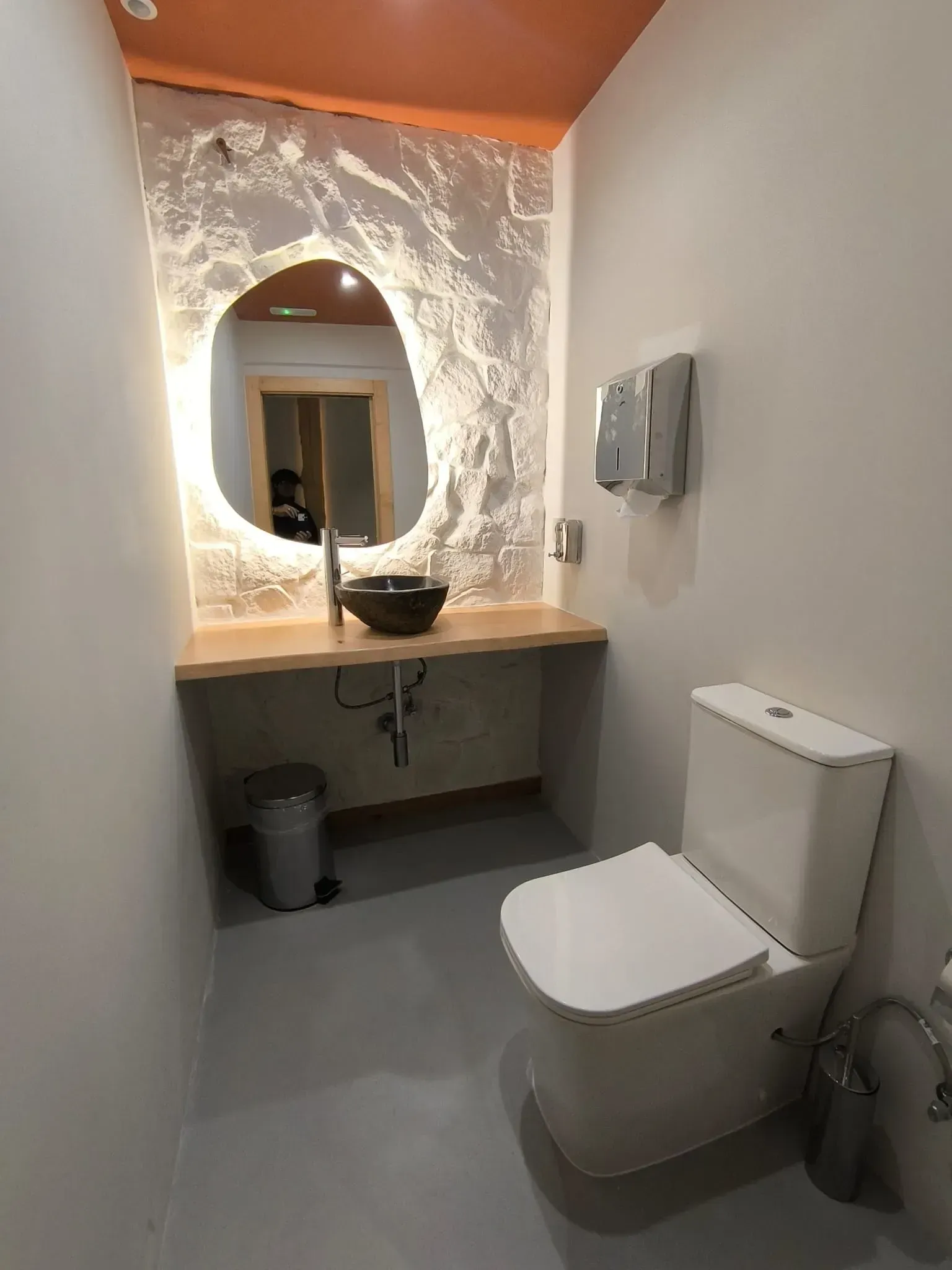 Baño moderno con inodoro, lavabo y espejo con marco iluminado. Combinación de colores gris, blanco y naranja.