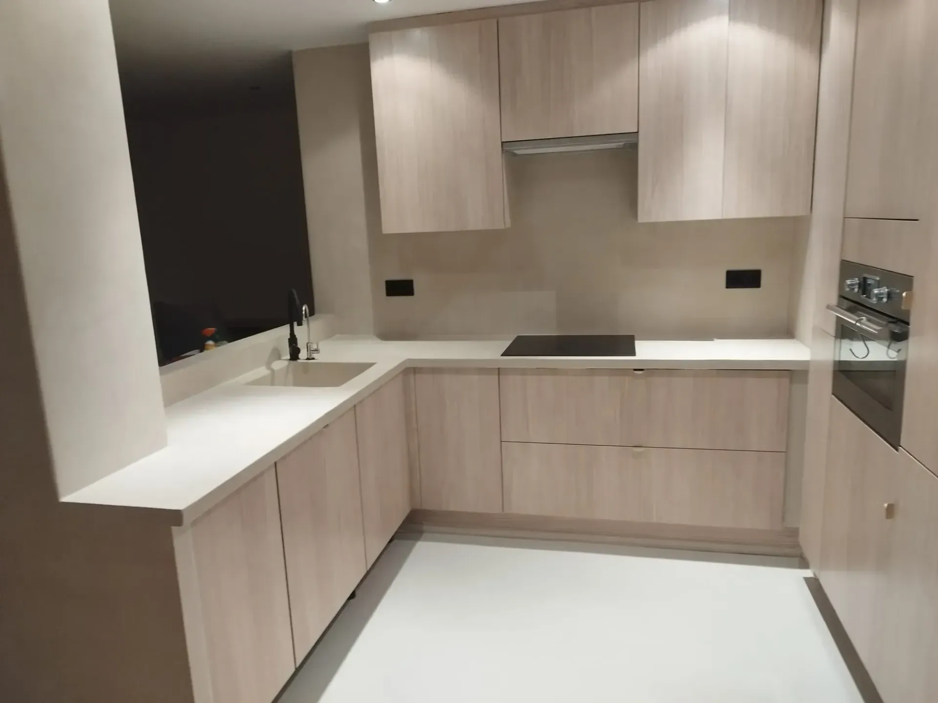 Cocina moderna con gabinetes de madera clara, encimeras blancas y piso blanco.