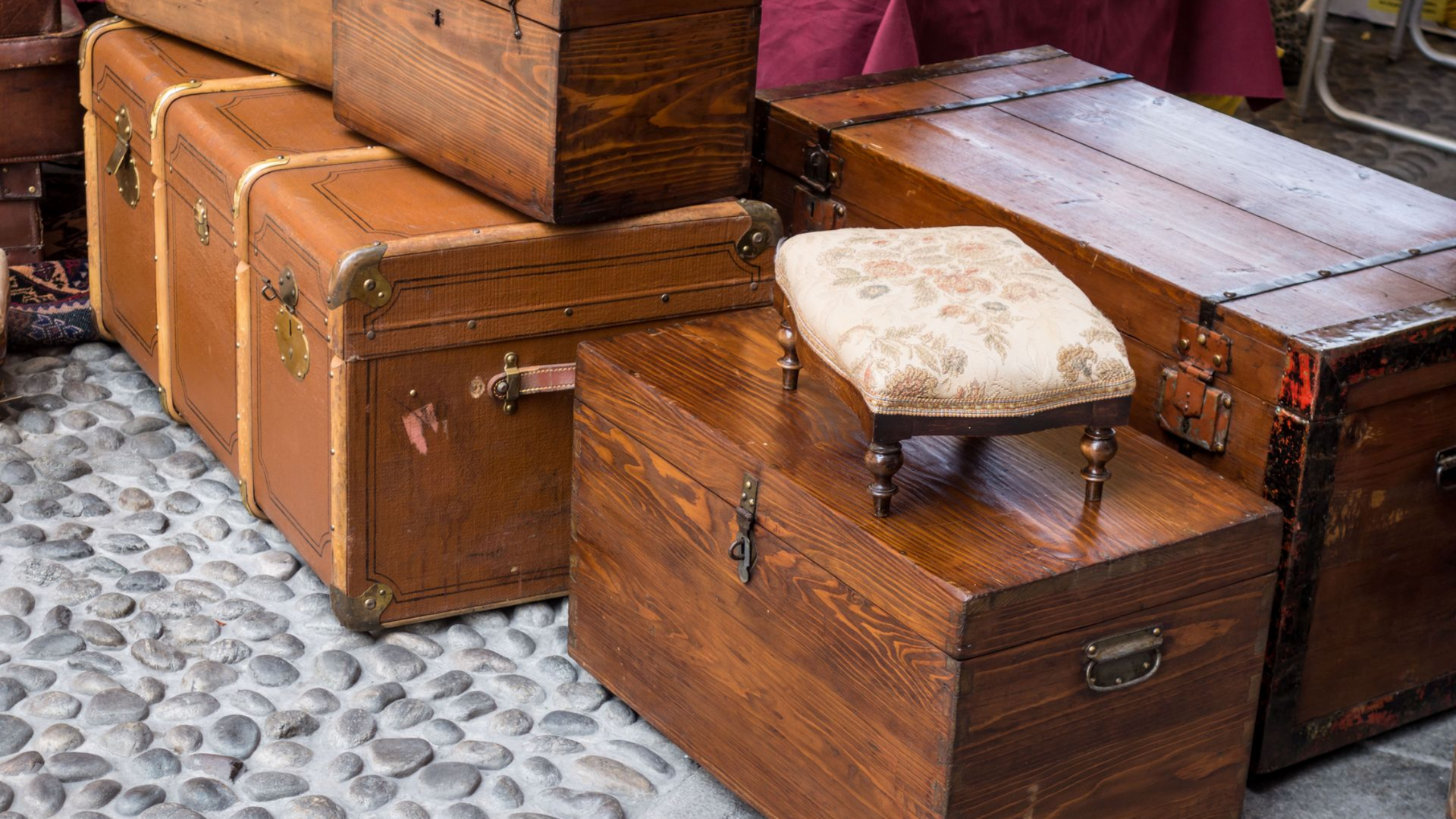 Malles en bois empilées, surmontées d'un petit repose-pieds rembourré. Sol pavé.
