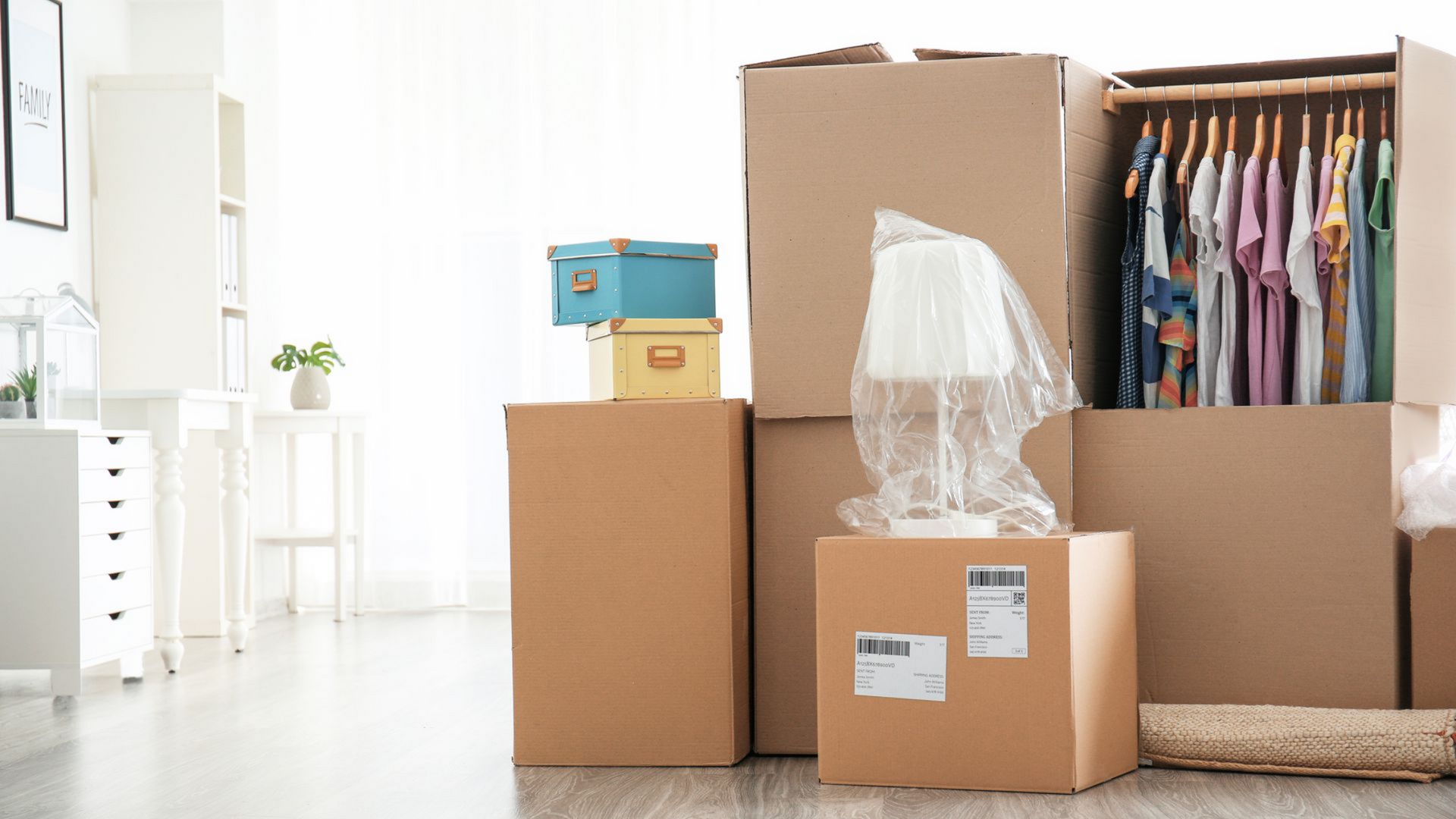 Des cartons empilés dans une pièce, des vêtements dans un placard ouvert, une lampe enveloppée dans du papier bulle.