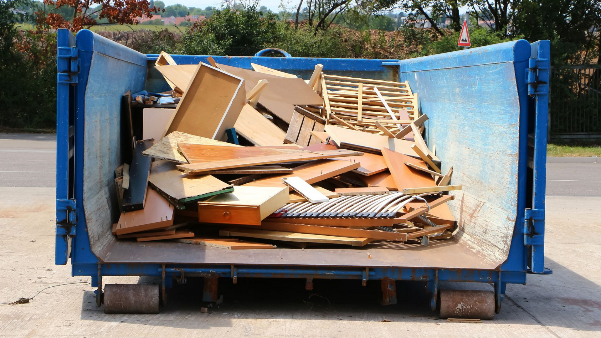 Benne bleue remplie de bois et de meubles jetés au bord d'une route.