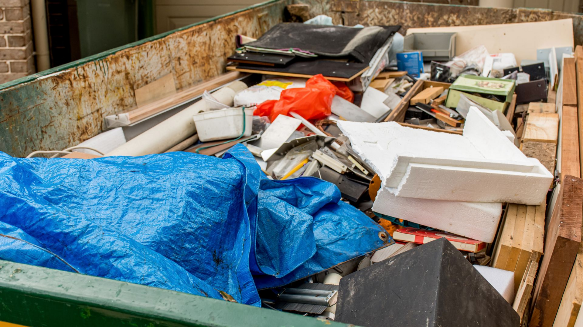 Conteneur rempli de débris de construction, notamment de bois, de plastique et d'une bâche bleue.