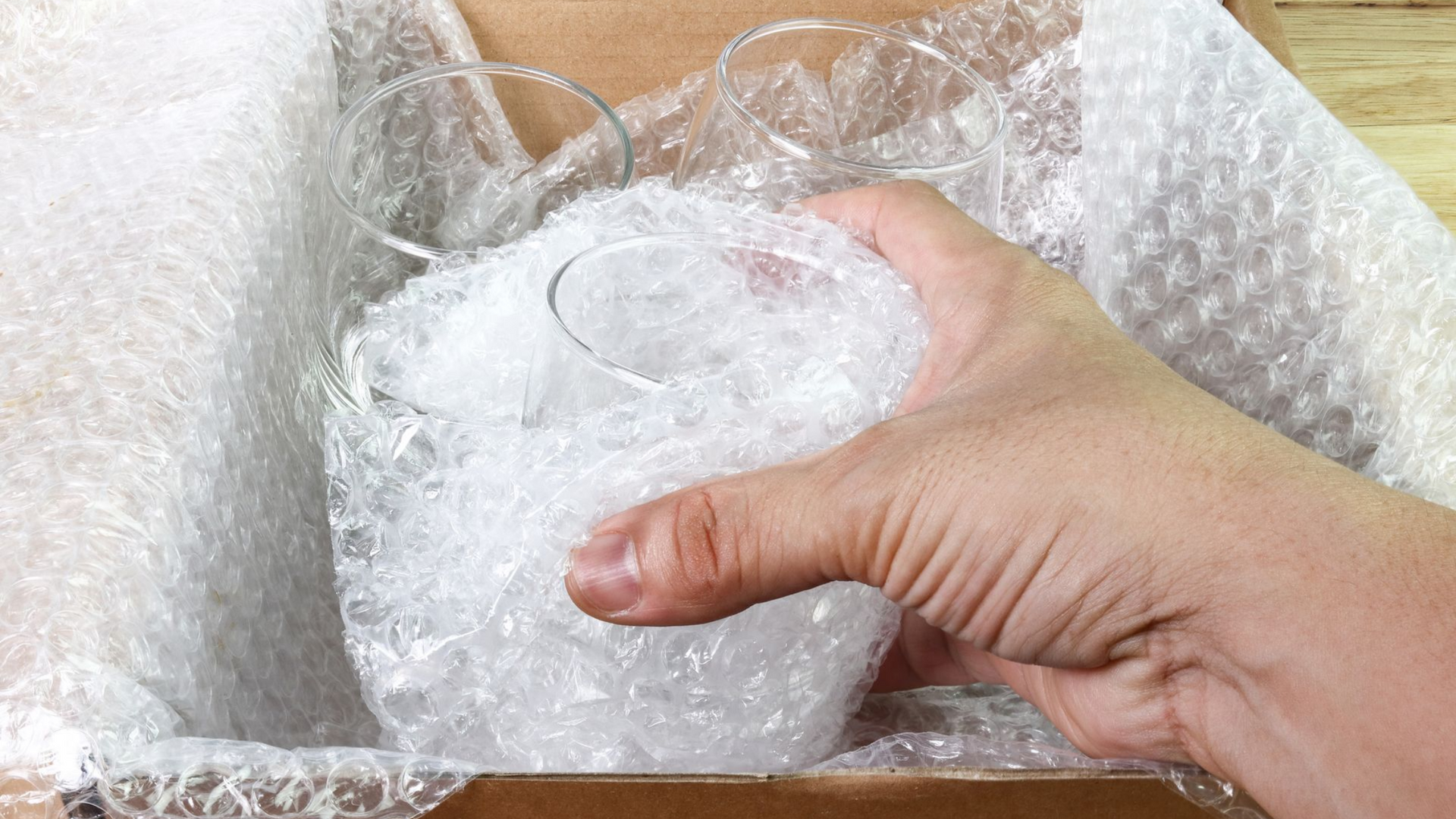 Main tenant un verre emballé dans du papier bulle dans une boîte en carton, entouré de plus de papier bulle.