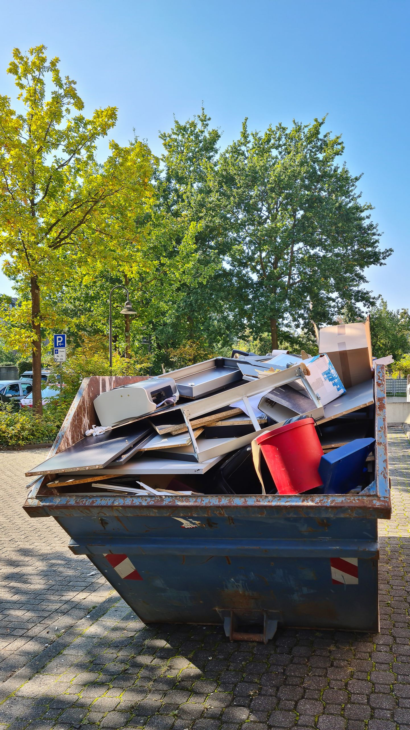 Benne bleue débordant de débris mélangés, avec des arbres et un ciel clair en arrière-plan.