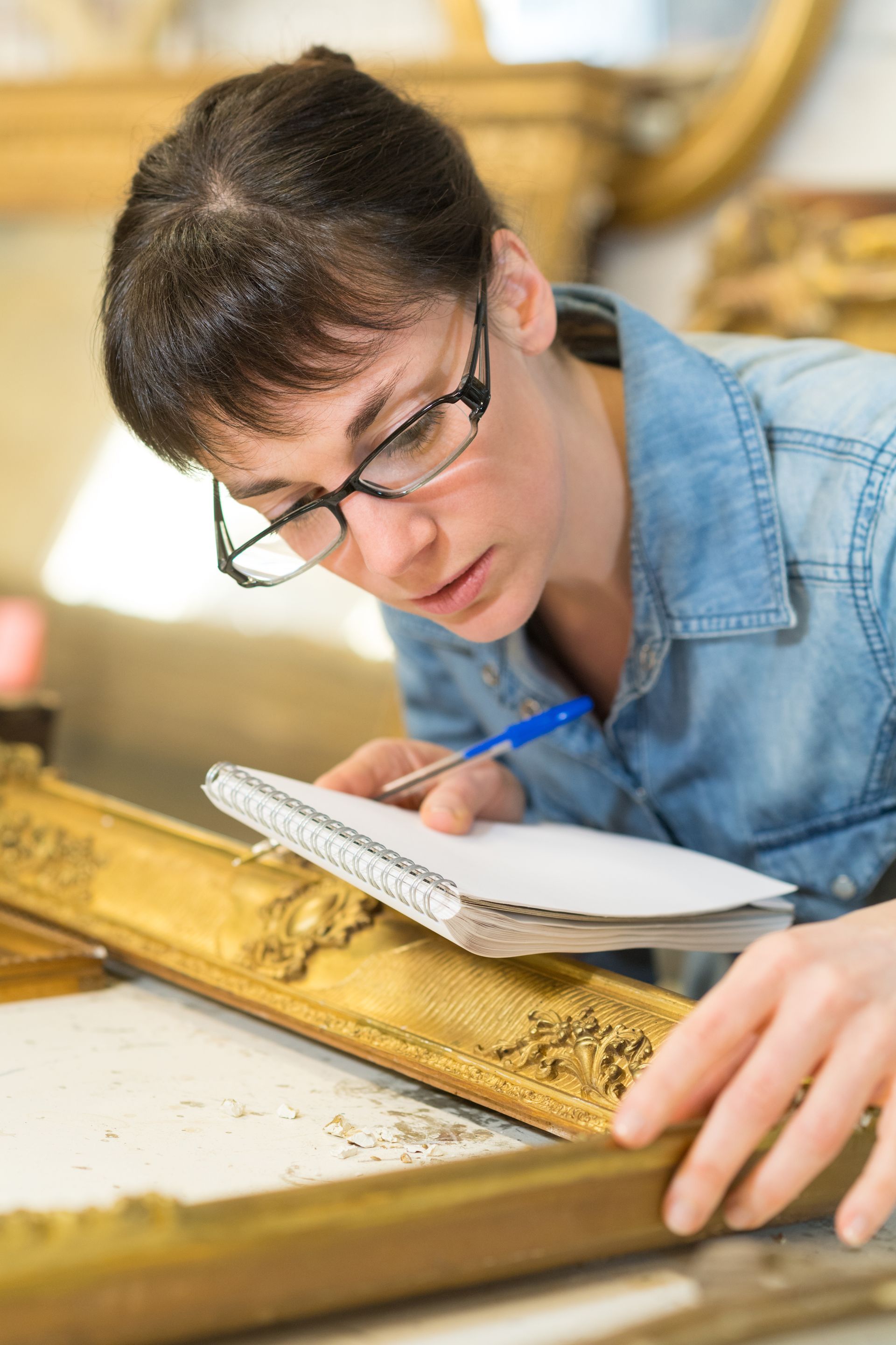 Une femme portant des lunettes examine une monture dorée ornée, prenant des notes avec un stylo dans un bloc-notes.