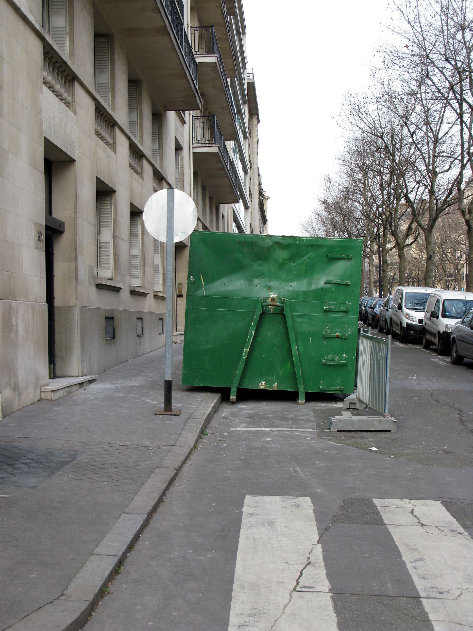 Une benne verte bloque un trottoir, avec un passage pour piétons au premier plan et un bâtiment sur la gauche.