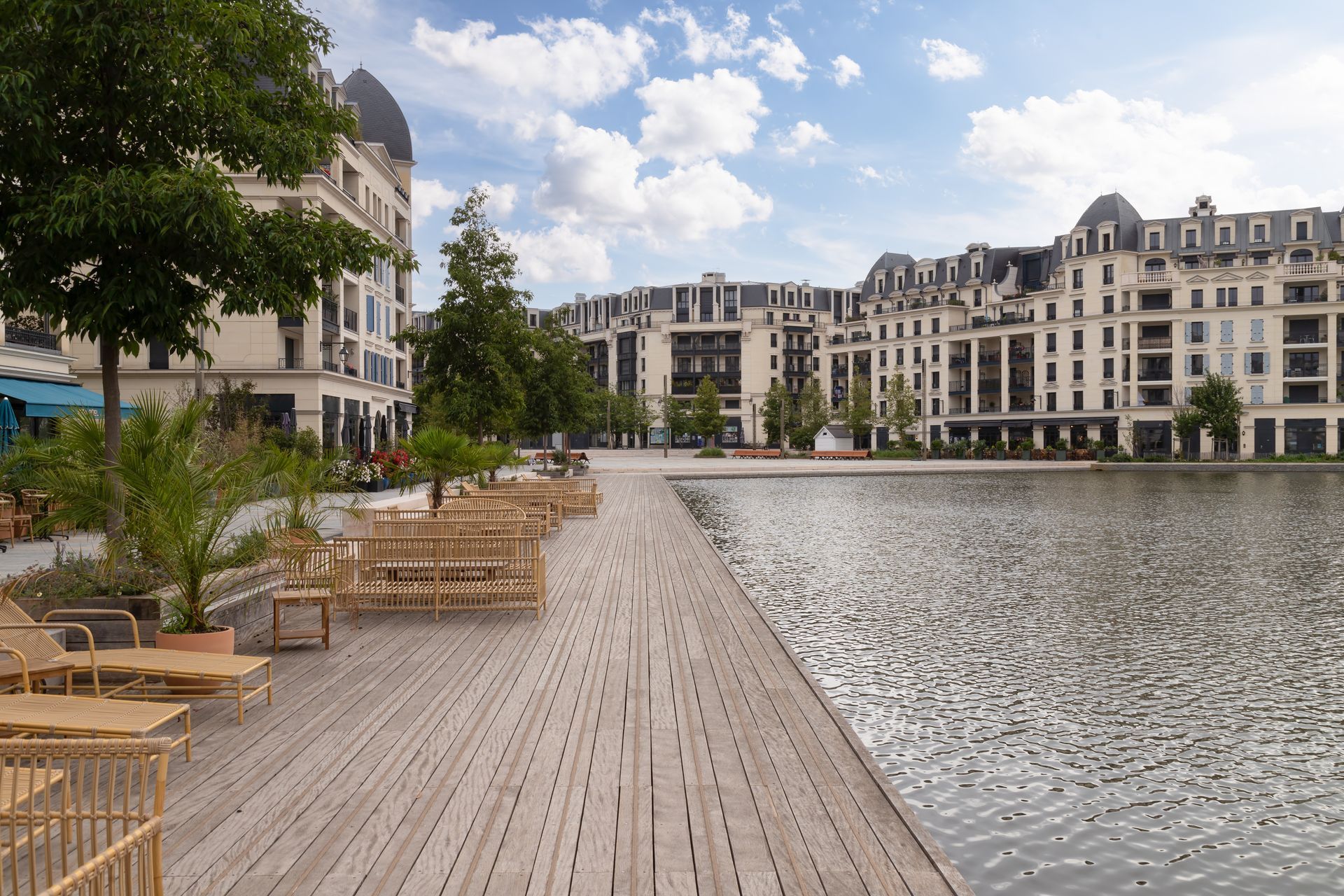 Promenade en bois au bord d'un lac, avec des bâtiments en arrière-plan. On aperçoit le ciel bleu, des bancs et des arbres.