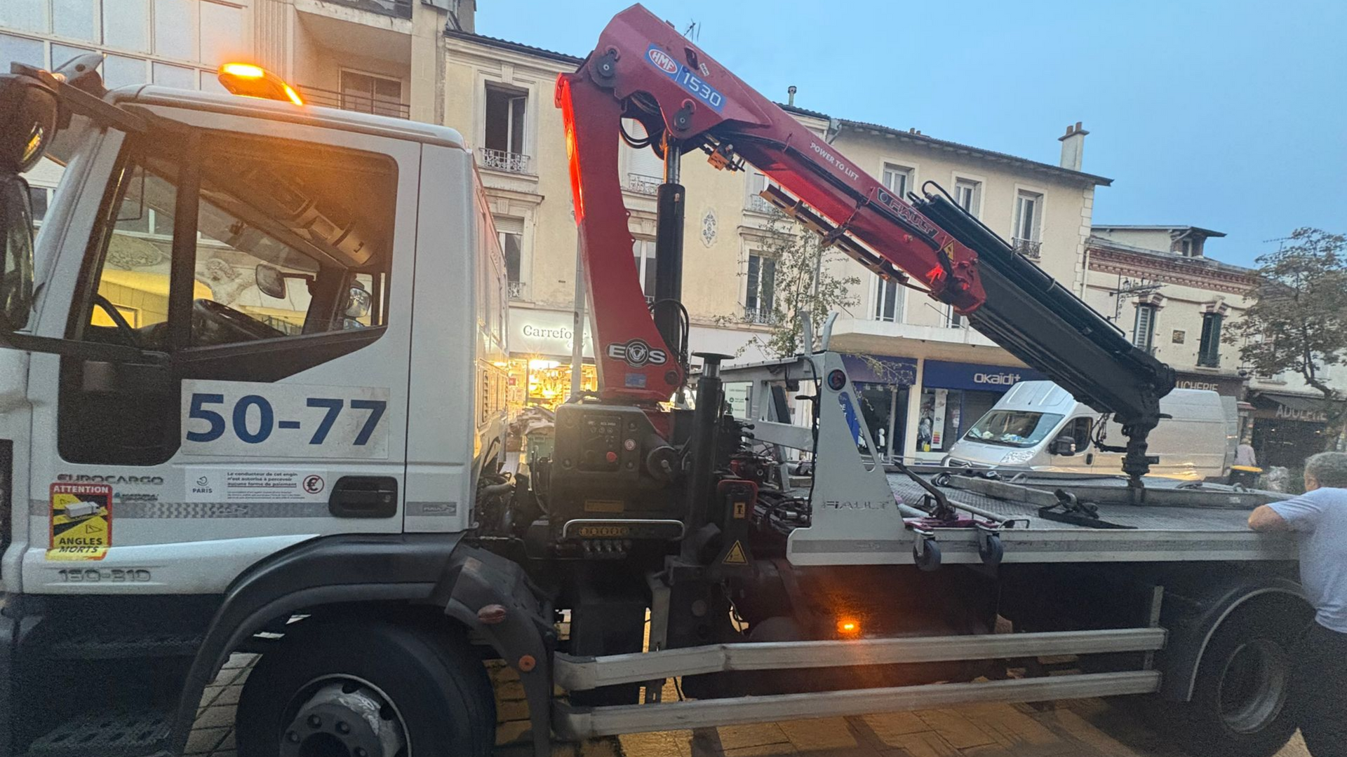 Dépanneuse avec grue sur la rue, feux allumés, remorquant une camionnette blanche.